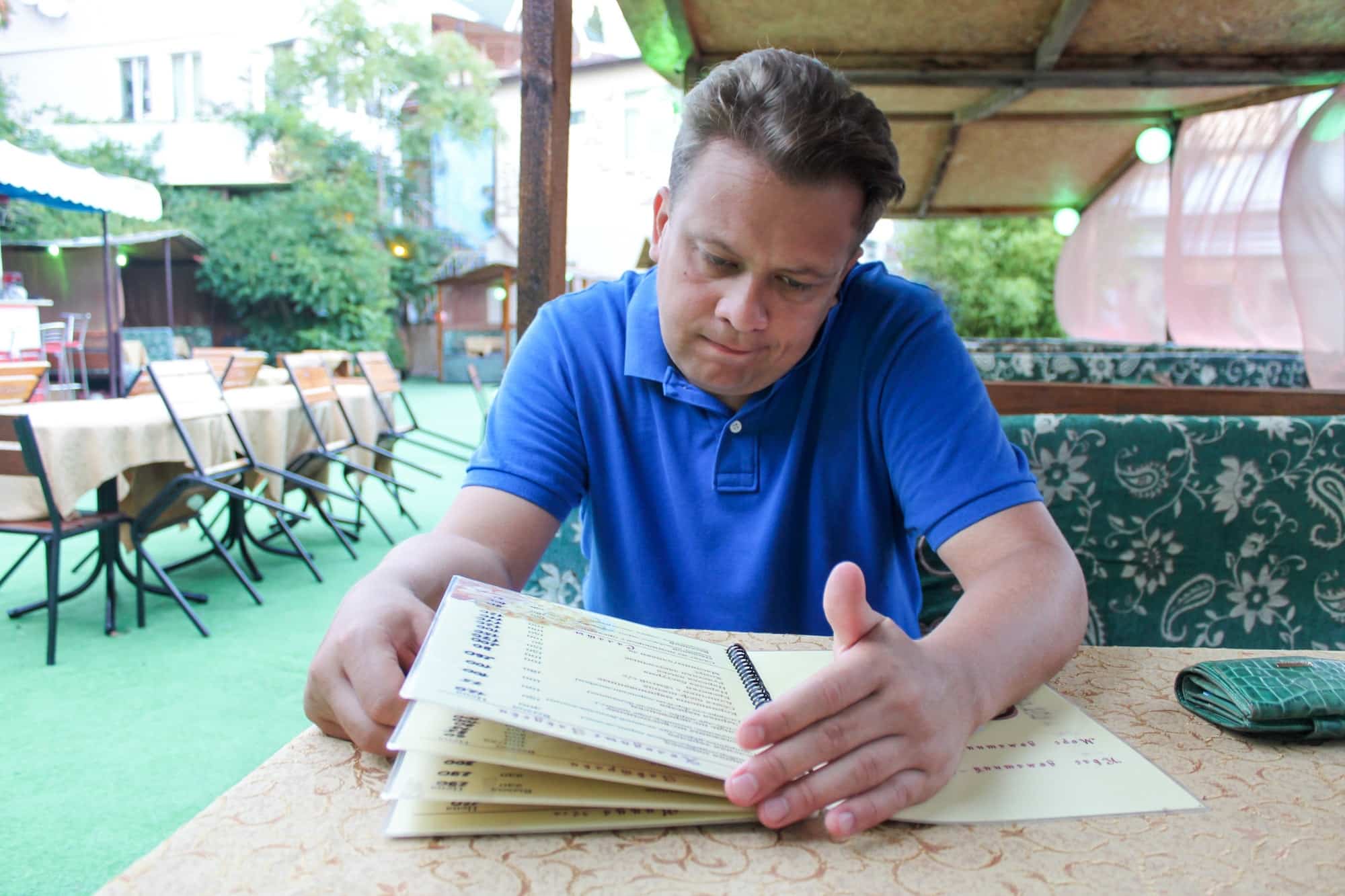 A young man the veranda of a summer restaurant looks at the menu. He is clearly confused by prices