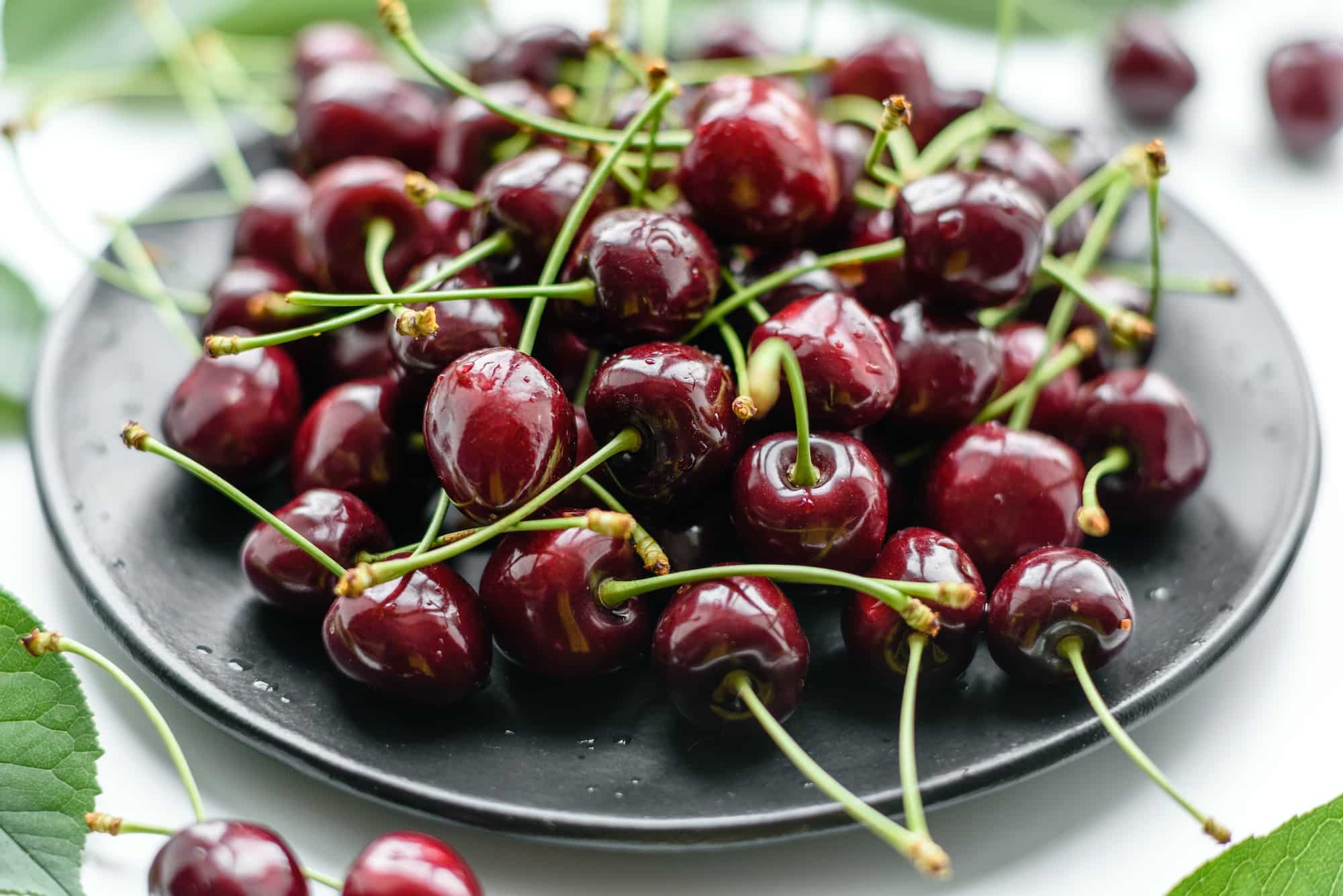 Fresh sweet cherries plate with leaves in water drops on stone background, top view