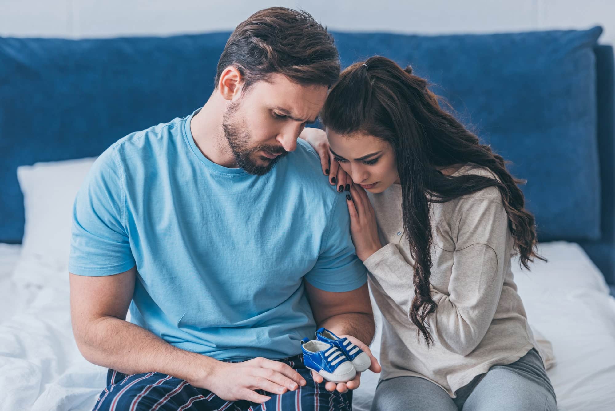 selective focus of grieving parents sitting on bed and holding baby shoes at home